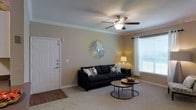 This single-story space presents a welcoming living room that balances comfort and style. The room features neutral-toned walls complemented by crisp crown molding that adds a refined architectural detail. A textured ceiling hosts a ceiling fan with an integrated light fixture, providing both air circulation and ambient lighting. A large window dressed with white curtains and horizontal blinds allows natural light to fill the room, creating a bright and airy atmosphere. The flooring is covered in plush, neutral-toned carpet, adding warmth underfoot. The front entryway is marked by a solid white door with a deadbolt, transitioning from a small hardwood-floored foyer into the carpeted living area. Wall décor includes a striking sunburst mirror that serves as a focal point on the main wall. The living space is designed to accommodate comfortable seating arrangements, and its overall layout offers a functional yet inviting environment. The warm, neutral color palette throughout the room contributes to a cozy and cohesive aesthetic, making the space feel welcoming from the moment you enter.
