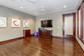 This single-floor fitness and wellness studio offers a thoughtfully designed open space ideal for yoga, movement classes, or group training sessions. The studio is characterized by rich, dark hardwood flooring that extends throughout the entire space, reflecting natural light and lending warmth to the cool gray walls. Recessed lighting and dual ceiling fans provide even illumination and comfortable airflow across the open floor area.

Large windows fitted with blinds line one wall, allowing natural light to filter in while maintaining privacy. Dark wood-framed glass doors provide access to an adjoining space, offering flexible layout options. A wall-mounted flat-screen television is positioned above a dark wood storage cabinet, providing a focal point for instructor-led sessions and media-based programming. Storage accessories, yoga blocks, and exercise mats are neatly kept on and around the cabinet, indicating full operational readiness.

Three vibrant circular abstract artworks adorn one wall, adding a touch of color and creative energy to the otherwise minimalist aesthetic. The overall ambiance is clean, open, and welcoming — balancing functional simplicity with a calm, professional atmosphere well-suited for wellness-focused activities.