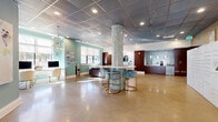 Located at 12751 Fair Lakes Circle in Fairfax, Virginia, this well-appointed leasing office serves as the welcoming hub of a residential community. The single-floor space boasts a spacious, open-concept design with polished concrete flooring throughout, lending a sleek, contemporary feel. A distinctive mosaic-tiled central column anchors the main area, surrounded by a circular bar-height table with gold-framed seating — an eye-catching focal point.

The lounge area is furnished with a plush sectional sofa, tufted ottoman, and accent chairs arranged around a flat-screen TV, creating a comfortable and inviting atmosphere for prospective and current residents alike. Soft teal-green accent walls, curated artwork, and layered lighting — including a statement drum pendant, sputnik chandelier, and recessed ceiling fixtures — contribute to the space's modern, upscale character.

A dedicated workstation area with monitors offers residents a convenient workspace. The back of the office features rich wood cabinetry housing a refreshment station with a coffee bar and refrigerator. A bank of smart package lockers and a separate mailroom with rows of individual mail slots provide practical resident services. The glass-paneled entrance foyer offers clear sightlines to the exterior, filling the space with natural light.