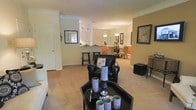 This single-floor residence showcases an open-concept floor plan that seamlessly connects the living, dining, and kitchen areas into one cohesive and functional space. The living area is anchored by plush, neutral-toned carpeting and features stylish black leather seating paired with a sleek black coffee table adorned with decorative crystal candle holders, lending the space a contemporary, polished feel. Crown molding runs throughout the interior, adding a refined architectural detail that elevates the overall design.

The kitchen is partially visible beyond a breakfast bar fitted with tall bar stools, offering a casual dining option in addition to the formal dining area beyond. The dining space is illuminated by an elegant chandelier, creating a warm and inviting atmosphere for gatherings. A second pendant light fixture is visible near the kitchen, reinforcing the layered lighting design throughout the home.

Framed artwork adorns the walls, contributing to a curated and thoughtfully decorated interior. The warm, earthy wall tones complement the neutral flooring and dark furniture accents, creating a balanced and welcoming ambiance across the entire living space.