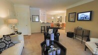 This single-floor residence showcases an open-concept floor plan that seamlessly connects the living, dining, and kitchen areas into one cohesive and functional space. The living area is anchored by plush, neutral-toned carpeting and features stylish black leather seating paired with a sleek black coffee table adorned with decorative crystal candle holders, lending the space a contemporary, polished feel. Crown molding runs throughout the interior, adding a refined architectural detail that elevates the overall design.

The kitchen is partially visible beyond a breakfast bar fitted with tall bar stools, offering a casual dining option in addition to the formal dining area beyond. The dining space is illuminated by an elegant chandelier, creating a warm and inviting atmosphere for gatherings. A second pendant light fixture is visible near the kitchen, reinforcing the layered lighting design throughout the home.

Framed artwork adorns the walls, contributing to a curated and thoughtfully decorated interior. The warm, earthy wall tones complement the neutral flooring and dark furniture accents, creating a balanced and welcoming ambiance across the entire living space.