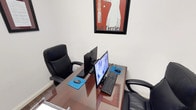 This single-floor office space offers a clean and functional environment designed for efficient, focused work. The layout centers around a sleek glass-topped desk that accommodates a dual-monitor computer setup, complete with keyboards and mouse pads, making it well-suited for tasks requiring multitasking or extended screen use. Two black ergonomic office chairs flank the workstation, providing comfortable seating for individual use or collaborative work sessions. The walls are painted in a neutral, light tone that contributes to a bright and calm atmosphere, while a framed Bull Terrier graphic print adds a touch of personality and visual interest to the space. The overall aesthetic is minimalist and professional, with clean lines and an uncluttered layout that promotes concentration. The compact scale of the space makes it ideal as a private office or a dedicated workstation within a larger commercial setting. Natural simplicity in the design keeps the focus on functionality, making this a practical and well-appointed workspace for day-to-day professional use.