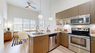 This single-story unit showcases a thoughtfully designed open-concept layout with notably high ceilings that create an airy, expansive atmosphere throughout. Light hardwood-style flooring flows seamlessly across the main living areas, enhancing the sense of continuity and space.

The kitchen is a standout feature, boasting warm wood-toned cabinetry that extends toward the tall ceilings, granite countertops, and a full suite of stainless steel appliances including a refrigerator, electric range, built-in microwave, and dishwasher. A peninsula with seating for two is illuminated by a globe pendant light, making it an ideal casual dining spot. Large windows allow natural light to fill the space, while a ceiling fan overhead ensures comfortable airflow.

The bedroom offers a relaxed retreat with tall ceilings, a wide window overlooking outdoor greenery, and a two-toned blue accent wall that adds depth and character. Carpet flooring provides warmth underfoot.

The bathroom features a generous vanity with granite countertops, warm wood cabinetry, a large mirror, and a tub-shower combination with tiled surround, maintaining the cohesive modern aesthetic found throughout the unit.