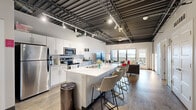 Located at 440 Des Moines St in the heart of downtown Des Moines, this two-bedroom, two-bathroom loft-style residence combines industrial character with modern finishes. Soaring exposed ceilings with steel beams and track lighting set a dramatic tone throughout the open-concept living area. The kitchen is a standout feature, equipped with stainless steel appliances, white shaker cabinetry, subway tile backsplash, and a generous island with bar seating — ideal for both cooking and entertaining. The kitchen flows seamlessly into the living room, where floor-to-ceiling windows and a glass balcony door frame sweeping views of the Des Moines skyline, flooding the space with natural light. A decorative "Des Moines" marquee sign adds a distinctive local touch to the living area. The primary bedroom offers direct balcony access with city views, a spacious closet, and wood-look flooring. The second bedroom features exposed ceiling details consistent with the loft aesthetic. Both bathrooms are finished with clean white vanities and wood-look tile flooring. Throughout the residence, light hardwood-style floors tie the spaces together, creating a cohesive, contemporary aesthetic that reflects the urban energy of downtown Des Moines.