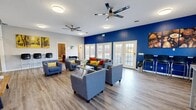 Located at 1095 Spruce St in Terre Haute, Indiana, this single-story clubhouse offers a thoughtfully designed community gathering space. The centerpiece is a generously sized open lounge area featuring wood-look flooring, dual ceiling fans, and a bold royal blue accent wall that adds a vibrant, modern energy to the space. Multiple sets of French doors line the exterior wall, flooding the room with natural light and providing access to the outdoors. Comfortable gray upholstered seating arranged throughout the lounge creates an inviting atmosphere for socializing, complemented by a wall-mounted flat-screen TV and casual game amenities. A bar-height counter with seating lines one wall, offering an additional gathering spot. The fully equipped kitchen features white cabinetry with stone-look countertops, a stainless steel sink, refrigerator, microwave, and a dining table with bar seating — all set against another striking blue accent wall. A dedicated exercise room provides space for fitness activities, while a private office supports administrative needs. Two well-finished bathrooms include rich wood-toned vanities, white countertops, and large mirrors with overhead lighting. Consistent wood-look flooring and a neutral gray and white color palette tie the entire clubhouse together cohesively.