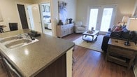 This single-story residence offers an open-concept layout that effortlessly connects the kitchen, dining, and living areas into one cohesive and functional space. The kitchen features a generous granite-style countertop island with bar seating, providing ample workspace and a natural gathering point that overlooks the living area. Rich hardwood-style flooring runs throughout, lending warmth and continuity to the space.

The living area is bright and welcoming, anchored by a soft area rug and centered around a coffee table with an industrial aesthetic. A patterned accent chair and a sideboard with distressed wood detailing add character and charm. Large French doors allow an abundance of natural light to pour into the space while offering direct access to the outdoors, enhancing the sense of openness.

Visible through a hallway, the bedroom is well-appointed and benefits from the same warm tones and natural light that define the rest of the home. Neutral wall colors throughout create a calm, versatile backdrop suited to a variety of décor styles. Overall, the space balances practicality with comfort, making it ideal for both everyday living and casual entertaining.