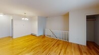 This 2-bedroom, 1-bathroom unit in Parsippany, NJ offers a practical and well-organized single-floor layout. Warm hardwood flooring runs throughout the main living areas and bedrooms, lending a consistent and inviting character to the space.

The living area is generously sized and features a large sliding glass door fitted with vertical blinds, allowing natural light to fill the room while providing access to an outdoor view with greenery. A wall-mounted air conditioning unit and baseboard heating ensure year-round comfort. A chandelier accent light adds a modest decorative touch near the dining area, which is positioned alongside a staircase with a white-railed landing visible from the main level.

The kitchen is equipped with warm oak cabinetry, dark laminate countertops, and a full suite of black appliances including a gas range, built-in microwave, dishwasher, and refrigerator. Light tile flooring grounds the kitchen area.

Both bedrooms feature the same hardwood flooring, neutral white walls, and windows with blinds that admit natural light. Each room includes baseboard heating, and one bedroom features a wall-mounted air conditioner.

The bathroom is clean and straightforward, offering a tiled tub-shower combination with a window above for natural ventilation and light.