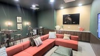 This single-floor lounge space is designed with a contemporary aesthetic that balances comfort and style. Deep forest green walls create a rich, immersive atmosphere, complemented by warm recessed lighting that adds a welcoming glow throughout the room. A large coral-toned sectional sofa serves as the centerpiece, offering generous seating and adorned with an assortment of textured throw pillows for a cozy, lived-in feel. Floor cushions provide additional flexible seating options for casual gatherings. Anchoring one wall is a wall-mounted flat-screen television set above a sleek built-in credenza featuring dark wood cabinetry and a light granite or stone countertop, which provides both storage and display space. A neutral accent wall frames this media area, creating a subtle contrast against the bold green tones. To the side, a compact high-top table with industrial-style bar stools offers a more upright seating arrangement, perfect for conversation or casual dining. The space is finished with light-toned flooring that brightens the room and complements the overall color palette. The layout and furnishings make this lounge well-suited for both relaxed recreation and social gatherings.