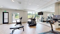 This modern fitness center offers a thoughtfully designed, single-level workout space with a clean, contemporary aesthetic. Light hardwood flooring runs throughout, complemented by a drop-panel ceiling with recessed lighting that keeps the environment bright and inviting.

The cardio zone is well-appointed with multiple TRUE-brand treadmills, elliptical trainers, and a stationary bike, alongside a flat-screen TV mounted on a full-length mirrored wall — a feature that visually expands the space and allows exercisers to monitor their form. Floor-to-ceiling windows line one wall, flooding the room with natural light and offering open views of a lush green outdoor landscape, creating an energizing workout environment.

The strength training area includes a multi-function cable machine suitable for a wide range of exercises, a dumbbell rack stocked with an extensive set of free weights, and a weight bench. Open wooden shelving provides organized storage for additional fitness accessories.

Black-framed glass doors add a stylish industrial touch and connect to adjacent areas, enhancing both accessibility and design continuity. An emergency exit with clear signage ensures safety throughout the space. Overall, the fitness center balances functionality with an upscale, airy atmosphere ideal for a focused and comfortable workout experience.