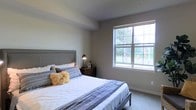 This single-floor residence offers a clean, modern aesthetic defined by neutral tones and thoughtful design details. The bedroom serves as a standout feature, showcasing a large multi-pane window that floods the room with natural light and offers a pleasant view of the surrounding greenery. The space is finished with light-toned carpet flooring and smooth walls in a soft gray palette, lending the room a calm and restful ambiance. A generously sized bed with an upholstered headboard anchors the room, complemented by a dark wood nightstand and a sleek adjustable reading lamp, creating a balanced blend of comfort and function. The window's bright outlook brings a sense of the outdoors inside, enhancing the overall airiness of the space. The room features clean lines and a minimalist approach to decor, with subtle textural accents introduced through layered bedding and decorative pillows. The ceiling is smooth and unadorned, contributing to the understated, contemporary feel of the space. Overall, this residence presents a serene and practical living environment well-suited to everyday comfort.