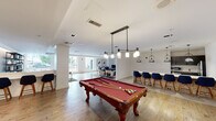 Located on Georgia Avenue in Silver Spring, Maryland, this single-floor community amenity space is thoughtfully designed to offer a variety of social and recreational experiences. The open-concept layout flows seamlessly between distinct activity zones, all unified by light hardwood flooring and crisp white walls that create a bright, welcoming atmosphere.

The game room area is anchored by a classic billiard table with a rich red felt surface, suspended beneath a stylish multi-pendant light fixture. Adjacent to it, a bar-height counter with navy blue upholstered stools provides a casual seating area, illuminated by coordinating pendant lights—ideal for conversation or co-working.

The lounge areas are furnished with an eclectic yet cohesive mix of seating, including tufted caramel leather sofas, curved armchairs in muted tones, and a large-screen wall-mounted television, all arranged atop boldly patterned area rugs. A built-in shelving unit with warm wood accents and decorative objects adds sophistication to one of the seating nooks, while abstract framed artwork enhances the curated aesthetic throughout.

A compact kitchenette, finished with dark cabinetry, white countertops, a stainless steel sink, microwave, mini-refrigerator, and coffee maker, supports the space's function as a gathering hub. Natural light filters in through windows, further enhancing the open, airy character of this well-appointed community retreat.