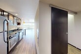 This single-floor unit offers a well-designed layout that balances open living spaces with practical, functional rooms. The combined kitchen and living area serves as the heart of the unit, where a galley-style kitchen features dark granite countertops, warm wood cabinetry, stainless steel appliances including a refrigerator, range, and dishwasher, and a double-basin sink. The kitchen flows seamlessly into the living area, which is brightened by expansive floor-to-ceiling windows offering views of the surrounding cityscape. Light wood-tone flooring runs throughout the main living spaces, complementing the clean white walls and modern finishes.

A hallway connects the main living area to the private spaces of the unit. A modern sliding barn-style door in dark wood with a sleek metal track adds a contemporary architectural detail to the corridor. The walk-in closet provides generous storage with wall-mounted wire shelving and hanging rods. The bathroom is spacious and accessible, featuring a soaking tub with a handheld showerhead, grab bars, and light stone-tile flooring. A dedicated laundry room houses a full-size side-by-side washer and dryer. Overall, the unit presents a clean, modern aesthetic throughout.