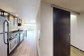 This single-floor unit offers a well-designed layout that balances open living spaces with practical, functional rooms. The combined kitchen and living area serves as the heart of the unit, where a galley-style kitchen features dark granite countertops, warm wood cabinetry, stainless steel appliances including a refrigerator, range, and dishwasher, and a double-basin sink. The kitchen flows seamlessly into the living area, which is brightened by expansive floor-to-ceiling windows offering views of the surrounding cityscape. Light wood-tone flooring runs throughout the main living spaces, complementing the clean white walls and modern finishes.

A hallway connects the main living area to the private spaces of the unit. A modern sliding barn-style door in dark wood with a sleek metal track adds a contemporary architectural detail to the corridor. The walk-in closet provides generous storage with wall-mounted wire shelving and hanging rods. The bathroom is spacious and accessible, featuring a soaking tub with a handheld showerhead, grab bars, and light stone-tile flooring. A dedicated laundry room houses a full-size side-by-side washer and dryer. Overall, the unit presents a clean, modern aesthetic throughout.