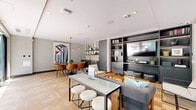 Located at 8377 Blackburn Ave in the heart of Los Angeles, this single-floor combined living and dining space delivers a refined, contemporary aesthetic with thoughtful design throughout. The room is anchored by a herringbone-patterned wood-look tile floor that adds warmth and visual interest across the entire space.

The living area features a built-in wall unit finished in dark charcoal, housing a large flat-screen television alongside open shelving and closed cabinetry — offering both display and storage functionality. A plush sofa paired with a slatted wood coffee table creates a comfortable lounge setting, while floor-to-ceiling glass sliding doors and curtain-wall windows flood the space with natural light and provide views to a lush outdoor terrace.

Transitioning seamlessly into the dining zone, a dark oval table surrounded by cognac leather chairs serves as the focal point, accented by a sculptural multi-bulb pendant light overhead. A striking large-scale abstract artwork adorns the textured accent wall, adding a bold artistic statement. Adjacent to the dining area, a compact kitchenette is outfitted with dark cabinetry, open industrial-style shelving, a sink, and an under-counter appliance — ideal for catering and entertaining. The clean white ceilings with recessed lighting complete the polished, modern ambiance of this well-appointed space.
