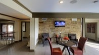 Located at 20 N Hill Parkway in Jackson, Mississippi, this single-floor space offers a thoughtfully designed interior that balances rustic charm with modern sensibility. The most commanding architectural feature is a full exposed stone accent wall, crafted from natural-cut masonry that lends warmth, texture, and a sense of permanence to the main living area. Light-toned wood-finish flooring runs throughout, providing a clean contrast to the rugged stonework and contributing to an open, airy atmosphere. The ceiling is finished in crisp white with recessed lighting, while dark olive crown molding frames the transition between walls and ceiling, adding a refined architectural detail. The living area is furnished with plush accent chairs and a round side table, creating an intimate seating arrangement well-suited for conversation or relaxation. A wall-mounted flat-screen television is centered on the stone wall, serving as a functional focal point. An open passageway connects the main seating area to an adjacent room, maintaining a sense of spatial flow throughout the layout. Ceiling-mounted speakers suggest an integrated audio system, enhancing the overall functionality of the space. Natural light filters in through windows visible in the connecting room, further brightening the interior. Overall, the space presents a welcoming and character-rich environment.