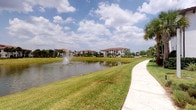 This single-story residence is located within a thoughtfully designed community that showcases distinct Mediterranean-inspired architecture. The buildings throughout the community feature characteristic terracotta tile rooflines, white stucco exteriors, and decorative detailing that lend a cohesive, upscale aesthetic to the surroundings.

The outdoor environment is a prominent feature of this property. A scenic pond serves as a central focal point of the community, complete with an active fountain that adds both visual interest and a calming ambiance. Well-maintained walking paths wind alongside the water's edge, bordered by manicured green lawns that invite leisurely strolls.

Tropical landscaping is found throughout the grounds, with tall, mature palm trees, ornamental shrubs, and carefully arranged plantings that reinforce the warm-weather, resort-like atmosphere. The lush greenery, combined with the open sky and waterfront views, creates a serene and welcoming outdoor setting.

The community's layout emphasizes walkability and open space, with pathways connecting buildings and garden areas. The combination of architectural consistency, water features, and tropical foliage contributes to a polished, peaceful environment suited to those who value both natural beauty and well-kept surroundings.