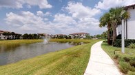 This single-story residence is located within a thoughtfully designed community that showcases distinct Mediterranean-inspired architecture. The buildings throughout the community feature characteristic terracotta tile rooflines, white stucco exteriors, and decorative detailing that lend a cohesive, upscale aesthetic to the surroundings.

The outdoor environment is a prominent feature of this property. A scenic pond serves as a central focal point of the community, complete with an active fountain that adds both visual interest and a calming ambiance. Well-maintained walking paths wind alongside the water's edge, bordered by manicured green lawns that invite leisurely strolls.

Tropical landscaping is found throughout the grounds, with tall, mature palm trees, ornamental shrubs, and carefully arranged plantings that reinforce the warm-weather, resort-like atmosphere. The lush greenery, combined with the open sky and waterfront views, creates a serene and welcoming outdoor setting.

The community's layout emphasizes walkability and open space, with pathways connecting buildings and garden areas. The combination of architectural consistency, water features, and tropical foliage contributes to a polished, peaceful environment suited to those who value both natural beauty and well-kept surroundings.