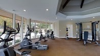 This fitness center offers a thoughtfully designed workout environment that combines functionality with an inviting atmosphere. The space is bathed in natural light, courtesy of large floor-to-ceiling windows that wrap along one wall, providing open views of the lush outdoor surroundings and creating an airy, energizing ambiance.

The interior features neutral, warm-toned walls and carpeted flooring, contributing to a comfortable and approachable setting. The ceiling incorporates architectural interest through recessed lighting and distinctive beamed or coffered details, adding a refined aesthetic to the utilitarian purpose of the space.

The facility is outfitted with a solid selection of cardio equipment, including elliptical trainers, upright and recumbent stationary bikes. A medicine ball rack stocked with a variety of weighted balls rounds out the functional training options, alongside additional strength and flexibility equipment positioned throughout the room. The layout is open and uncluttered, allowing for ease of movement between stations.

A private room or storage area is accessible through a door at the far end, adding to the overall utility of the space. Together, the abundant natural light, well-maintained equipment, and clean architectural design make this fitness center a practical and pleasant destination for daily exercise.