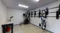 This dedicated bicycle storage room is designed with functionality and organization in mind. The space features a series of black wall-mounted bike racks installed along one wall, allowing multiple bicycles to be hung vertically or at angles, making efficient use of the available floor and wall space. The racks are numbered, suggesting an assigned or managed storage system, ideal for a shared residential or commercial building setting.

The room is finished with smooth, light gray painted walls and a concrete floor, offering a durable and low-maintenance environment suited for storing equipment. Overhead fluorescent lighting provides ample illumination throughout the space, ensuring visibility and ease of access at any time of day.

The layout accommodates a variety of bicycle types, including road bikes, mountain bikes, and children's bikes, demonstrating its versatility. Additional hooks and brackets extend along the wall to provide capacity for several more bicycles beyond those currently stored. The open floor area allows for easy maneuvering when retrieving or storing bikes. Overall, the room is a practical, well-organized solution for secure bicycle storage.