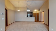 This single-floor residence showcases a thoughtfully designed open-concept layout that seamlessly connects the living area and kitchen. The living room is spacious and inviting, featuring vaulted ceilings that enhance the sense of height and openness. A ceiling fan is centrally mounted, adding both function and a casual aesthetic to the room. Soft, neutral-toned carpeting covers the living area floor, contributing to a warm and comfortable ambiance.

A decorative pendant light fixture hangs near the kitchen transition, serving as a design accent that bridges the two spaces. The kitchen is defined by a raised peninsula counter, which provides both a natural room divider and an area for casual dining or food preparation. Dark wood cabinetry lines the kitchen walls, complemented by stainless steel appliances and a countertop with subtle patterning. Recessed and fluorescent lighting illuminates the kitchen efficiently.

Beyond the kitchen, the space transitions into a hallway with hardwood-style flooring and warm wood-toned doors, hinting at additional rooms within the residence. The overall color palette is neutral and cohesive, with beige walls, brown trim, and earthy tones throughout, creating a grounded and welcoming atmosphere. The single-floor plan offers practical, accessible living in a well-appointed layout.