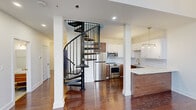 Located at 50 S Jefferson St in downtown Dayton, Ohio, this two-level unit offers a thoughtfully designed layout spread across a generous main floor and a smaller upper level. Rich hardwood floors flow throughout both levels, anchoring a cohesive and polished aesthetic.

The main level is centered around an open-plan living and kitchen area. The kitchen is well-appointed with stainless steel appliances including a range, microwave, and refrigerator, complemented by white subway tile backsplash, two-tone cabinetry in white and warm walnut, and a marble-look countertop with a gold-tone faucet. A multi-bulb brass pendant light adds a modern industrial touch. Soaring ceilings with recessed lighting and large black-framed windows allow ample natural light to fill the space. A bathroom on the main level features full subway tile surround, a sliding glass shower door, and brushed gold hardware throughout.

A striking black iron spiral staircase serves as both a functional and sculptural centerpiece, connecting the main level to the upper floor. Upstairs, the landing opens to multiple rooms featuring board-and-batten wainscoting, clerestory windows, angled architectural details, and brass sconce lighting. White doors with gold hardware carry the design language consistently through every space, creating a refined and cohesive urban residence.