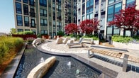The Zen Garden is a thoughtfully designed outdoor retreat set within the courtyard of a contemporary high-rise building. At its centerpiece is a curvilinear water feature that winds through the space, punctuated by large natural boulders and gentle fountains that create a calming acoustic backdrop. The water channel is bordered by a combination of irregular flagstone pathways and textured paver surfaces, guiding visitors through distinct seating areas.

A circular deck platform serves as a focal gathering point, furnished with teak-framed chaise lounges and low benches ideal for relaxation. Additional seating areas with Adirondack-style chairs are positioned among raised planting beds, which showcase vibrant Japanese maple trees with striking deep-red foliage, ornamental grasses, and lush perennial plantings that add vivid seasonal color.

A covered pavilion with dark structural columns and a warm wood-paneled ceiling anchors one end of the garden, offering a shaded transition space with outdoor dining furniture and floor-to-ceiling glass doors. The open-air layout receives abundant natural light throughout the day, while the surrounding building facades frame the space, creating a sheltered yet airy urban sanctuary designed for quiet contemplation and relaxation.
