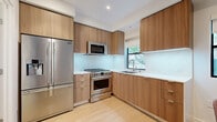 Located in the desirable Brentwood neighborhood of Los Angeles, this efficiently designed one-bedroom, one-bathroom unit offers a thoughtfully curated modern aesthetic across its single floor. The open kitchen serves as a standout feature, outfitted with full-height flat-front wood cabinetry in a warm walnut tone, white quartz countertops with under-cabinet lighting, and a full suite of stainless steel appliances including a gas range, built-in microwave, and a French door refrigerator. A window above the sink frames views of lush greenery, filling the kitchen with natural light. The space flows seamlessly into a casual dining area, reinforcing the open-concept layout. Light-toned hardwood flooring runs throughout, lending warmth and visual continuity to every room. The bedroom is a clean, minimal space with crisp white walls, a wide window overlooking foliage, and a wall-mounted television. The bathroom continues the modern design language, featuring a wood vanity with a white countertop, an integrated backlit mirror, and a tiled shower enclosure. Clean lines and a neutral palette define the overall ambiance, creating a polished and cohesive living environment.