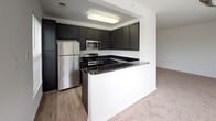 This single-floor residence offers a clean, functional layout designed for comfortable everyday living. The kitchen serves as a standout feature, equipped with dark cabinetry, granite-style dark countertops, stainless steel appliances including a refrigerator, gas range, and microwave, and a peninsula breakfast bar that opens to the adjacent living area. The flooring transitions from wood-look vinyl in the kitchen to plush neutral carpet throughout the living spaces and bedrooms, creating a warm and cohesive feel. The open living and dining areas are bathed in natural light, courtesy of multiple windows dressed with vertical blinds and horizontal blinds, offering views of lush greenery outside. The bedrooms are generously sized with soft carpeting, bright white walls, and large windows that continue the theme of natural illumination. The primary bedroom includes direct access to a private bathroom and a dedicated closet area. The bathrooms feature dark vanities with contrasting dark countertops, wood-look vinyl flooring, and clean white fixtures, including a tub-shower combination. The overall aesthetic is modern and neutral, providing a versatile canvas throughout the entire space.