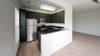 This single-floor residence offers a clean, functional layout designed for comfortable everyday living. The kitchen serves as a standout feature, equipped with dark cabinetry, granite-style dark countertops, stainless steel appliances including a refrigerator, gas range, and microwave, and a peninsula breakfast bar that opens to the adjacent living area. The flooring transitions from wood-look vinyl in the kitchen to plush neutral carpet throughout the living spaces and bedrooms, creating a warm and cohesive feel. The open living and dining areas are bathed in natural light, courtesy of multiple windows dressed with vertical blinds and horizontal blinds, offering views of lush greenery outside. The bedrooms are generously sized with soft carpeting, bright white walls, and large windows that continue the theme of natural illumination. The primary bedroom includes direct access to a private bathroom and a dedicated closet area. The bathrooms feature dark vanities with contrasting dark countertops, wood-look vinyl flooring, and clean white fixtures, including a tub-shower combination. The overall aesthetic is modern and neutral, providing a versatile canvas throughout the entire space.