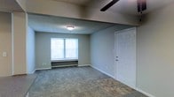 This single-floor space showcases a functional and straightforward layout designed for comfortable everyday living. The main living area is finished with wall-to-wall carpeting that transitions to hard flooring near the entryway, providing both comfort and practicality. A white paneled entry door serves as the main access point, complemented by clean white baseboards that run along the perimeter of the room.

The living area benefits from natural light through a centrally positioned window that frames views of surrounding greenery, creating a bright and airy atmosphere. Below the window, a baseboard heating unit ensures efficient climate control throughout the space. The textured ceiling adds a classic residential character to the room, while a ceiling fan overhead contributes to air circulation and comfort year-round.

The walls are finished in a neutral, muted tone that provides a versatile backdrop suitable for a variety of interior styles. Overall, the space presents a clean, move-in-ready environment with practical features well-suited for day-to-day living.