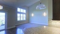 This two-story space showcases thoughtful architectural design and quality craftsmanship throughout. The main living and dining area immediately draws the eye upward with its vaulted ceiling, which amplifies the sense of openness and volume. A large multi-pane window floods the room with natural light, offering views of surrounding greenery and creating a bright, airy ambiance. The walls are finished in a soft, neutral tone that complements the overall palette of the space.

Underfoot, rich hardwood flooring transitions seamlessly into carpeted areas, delineating different functional zones while maintaining visual continuity. A striking glass pendant light hangs overhead, serving as a focal point and adding a touch of elegance. Two vertical wall sconces provide additional ambient lighting along the walls, enhancing the room's warm, inviting character.

A granite surface — likely a kitchen island or countertop — is partially visible in the foreground, hinting at high-end finishes carried throughout the space. The clean lines, quality materials, and carefully considered lighting combine to give this two-story property a polished yet comfortable atmosphere well-suited for everyday living and entertaining.