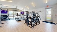 Located in Lakeville, Minnesota, this fitness center offers a comprehensive and thoughtfully designed workout environment within a compact yet efficiently utilized single-floor space. The open layout maximizes functionality, providing room for a diverse range of exercise equipment and activities.

The cardio zone is prominently stocked with Matrix-brand treadmills and elliptical trainers, allowing multiple users to train simultaneously. A Matrix upright cycle adds to the cardio options. For strength training, the facility includes a cable functional trainer, a dumbbell rack with an assorted weight selection, and a variety of stability and medicine balls to support dynamic and core-focused workouts. An exercise mat station is also available for stretching and floor exercises.

The interior design strikes a balance between energetic and clean. Light hardwood-style flooring runs throughout, complementing the bright white walls and modern linear LED lighting overhead. A standout feature is the exposed white brick accent wall adorned with a large-format Muhammad Ali quote — "Float like a butterfly, sting like a bee" — accompanied by oversized decorative boxing gloves, setting an inspiring and athletic tone. Large windows and a glass garage-style door invite natural light and provide views to the outdoors, enhancing the open, airy ambiance. A ceiling fan adds comfort during intense workouts. The space is well-ventilated, clean, and purpose-built for a focused fitness experience.