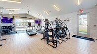 Located in Lakeville, Minnesota, this fitness center offers a comprehensive and thoughtfully designed workout environment within a compact yet efficiently utilized single-floor space. The open layout maximizes functionality, providing room for a diverse range of exercise equipment and activities.

The cardio zone is prominently stocked with Matrix-brand treadmills and elliptical trainers, allowing multiple users to train simultaneously. A Matrix upright cycle adds to the cardio options. For strength training, the facility includes a cable functional trainer, a dumbbell rack with an assorted weight selection, and a variety of stability and medicine balls to support dynamic and core-focused workouts. An exercise mat station is also available for stretching and floor exercises.

The interior design strikes a balance between energetic and clean. Light hardwood-style flooring runs throughout, complementing the bright white walls and modern linear LED lighting overhead. A standout feature is the exposed white brick accent wall adorned with a large-format Muhammad Ali quote — "Float like a butterfly, sting like a bee" — accompanied by oversized decorative boxing gloves, setting an inspiring and athletic tone. Large windows and a glass garage-style door invite natural light and provide views to the outdoors, enhancing the open, airy ambiance. A ceiling fan adds comfort during intense workouts. The space is well-ventilated, clean, and purpose-built for a focused fitness experience.