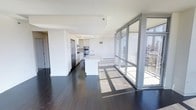 This single-floor high-rise unit offers a refined, open-concept living experience defined by clean lines and contemporary finishes throughout. The expansive living and kitchen area is anchored by a dramatic wall of floor-to-ceiling windows, flooding the interior with abundant natural light and framing impressive panoramic city views. Rich, dark hardwood flooring runs throughout the main living space, providing a striking contrast to the bright, white palette of the kitchen and ceilings.

The kitchen is thoughtfully designed with crisp white cabinetry, stainless steel appliances including a built-in refrigerator and double wall ovens, and a central island with a light stone countertop that serves as both a prep area and a casual gathering point. Track lighting above the kitchen ensures excellent task illumination, complementing the natural light that pours in from the adjacent windows.

The open layout creates a seamless flow between the kitchen and living areas, making the space feel expansive and airy. The large windows not only provide sweeping urban views but also open to what appears to be an outdoor terrace or balcony, further extending the living space and enhancing the connection to the surrounding cityscape. The overall ambiance is modern, bright, and sophisticated.