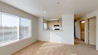 This single-floor residential unit offers a thoughtfully laid-out living space with a clean, contemporary design throughout. The open living area is carpeted and filled with natural light streaming through large windows fitted with horizontal blinds, creating a bright and welcoming atmosphere. A pass-through kitchen overlooks the living space, featuring light wood cabinetry, white countertops, a black electric range with a ventilation hood, and a window that brings in additional natural light — making the kitchen feel connected yet distinct from the main living area. The bedroom accommodates comfortable living with soft carpeting, a large window for natural light, and a spacious closet with sliding doors offering ample storage. A second room, also carpeted and windowed, provides flexible use as an additional bedroom or multipurpose space. The bathroom is well-appointed with warm wood-style flooring, a single-basin vanity with light cabinetry, a large mirror, and a full-sized soaking tub with a shower. Neutral tones and consistent finishes are carried throughout the unit, contributing to a cohesive and functional aesthetic. The overall layout, as seen in the dollhouse view, is efficiently organized to maximize usable space.