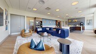 Located at 360 State St in New Haven, CT, this thoughtfully designed amenity lounge offers a generous open-plan layout that seamlessly blends comfort and sophistication. The space is anchored by a plush lounge area furnished with deep navy blue tufted sofas and warm tan leather swivel chairs, arranged around round coffee tables atop a soft area rug — creating an inviting social atmosphere. A striking blue textured accent wall serves as a focal point, complemented by warm gold wall sconces and rich wood built-in shelving that adds depth and warmth to the interior.

The kitchen and bar area is a standout feature, boasting an expansive white quartz waterfall-edge island illuminated by a cluster of bold brass pendant lights. Blue subway tile backsplash, dark wood cabinetry, and a row of sleek bar seating complete this refined entertaining zone. Herringbone-patterned flooring transitions between light and dark tones, visually defining the lounge and kitchen areas.

Floor-to-ceiling windows along one side of the space flood the interior with natural light while offering sweeping outdoor views of the surrounding landscape. Built-in window bench seating invites residents to relax and take in the scenery. The overall ambiance is polished yet welcoming, ideal for socializing or unwinding in style.
