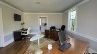 This single-floor office space combines classic architectural character with a practical, professional layout. The main workspace is generously sized, featuring rich hardwood flooring, crisp crown molding, and a textured ceiling that adds depth to the room. Soft, neutral gray walls create a calm and focused atmosphere throughout.

The room accommodates multiple workstations, including a large dark wood executive desk positioned near the center, equipped with computer monitors and office equipment, and a secondary desk along the perimeter, offering ample workspace for productivity. A decorative area rug anchors the central workspace, adding warmth and visual interest to the hardwood floors.

Natural light fills the space through large, multi-pane windows that frame views of the surrounding greenery, enhancing the open and airy feel of the room. A stylish pedestal side table adds a modern accent among the more traditional furnishings.

An interior doorway connects to an adjacent room, visible in the background, which features white cabinetry and additional space — suggesting a utility or kitchenette area. The overall ambiance of the space is professional yet welcoming, blending traditional design elements with functional office needs, making it well-suited for a private practice, small business, or executive office setting.