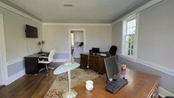 This single-floor office space combines classic architectural character with a practical, professional layout. The main workspace is generously sized, featuring rich hardwood flooring, crisp crown molding, and a textured ceiling that adds depth to the room. Soft, neutral gray walls create a calm and focused atmosphere throughout.

The room accommodates multiple workstations, including a large dark wood executive desk positioned near the center, equipped with computer monitors and office equipment, and a secondary desk along the perimeter, offering ample workspace for productivity. A decorative area rug anchors the central workspace, adding warmth and visual interest to the hardwood floors.

Natural light fills the space through large, multi-pane windows that frame views of the surrounding greenery, enhancing the open and airy feel of the room. A stylish pedestal side table adds a modern accent among the more traditional furnishings.

An interior doorway connects to an adjacent room, visible in the background, which features white cabinetry and additional space — suggesting a utility or kitchenette area. The overall ambiance of the space is professional yet welcoming, blending traditional design elements with functional office needs, making it well-suited for a private practice, small business, or executive office setting.