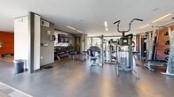 Located at 510 Taylor Place in Nashville, TN, this contemporary fitness center is thoughtfully designed to accommodate a full range of workout needs within a single, well-appointed floor. The facility is divided into distinct functional zones, each finished with purposeful materials that suit their use.

The cardio area features hardwood flooring and is lined with a wide array of equipment, including stationary bikes, ellipticals, and stair climbers, all positioned in front of expansive floor-to-ceiling windows that flood the space with natural light and provide views of the surrounding greenery and an outdoor pool area. A large wall-mounted mirror amplifies the sense of openness in this zone.

The strength and free weights area transitions to polished concrete flooring and is well-stocked with a comprehensive dumbbell rack, cable machines, and multi-use strength stations. A wall-mounted television adds convenience for users during workouts.

A dedicated open studio space offers hardwood flooring, full-length mirrors along one wall, and dramatic diagonal structural beams framed against the glass exterior wall—providing an architectural focal point alongside pool and courtyard views. Medicine balls and other functional training accessories are readily available in this area.

The entry zone features mixed carpet tile flooring and exposed brick walls, creating an industrial yet welcoming aesthetic that ties the overall design together.