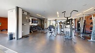 Located at 510 Taylor Place in Nashville, TN, this contemporary fitness center is thoughtfully designed to accommodate a full range of workout needs within a single, well-appointed floor. The facility is divided into distinct functional zones, each finished with purposeful materials that suit their use.

The cardio area features hardwood flooring and is lined with a wide array of equipment, including stationary bikes, ellipticals, and stair climbers, all positioned in front of expansive floor-to-ceiling windows that flood the space with natural light and provide views of the surrounding greenery and an outdoor pool area. A large wall-mounted mirror amplifies the sense of openness in this zone.

The strength and free weights area transitions to polished concrete flooring and is well-stocked with a comprehensive dumbbell rack, cable machines, and multi-use strength stations. A wall-mounted television adds convenience for users during workouts.

A dedicated open studio space offers hardwood flooring, full-length mirrors along one wall, and dramatic diagonal structural beams framed against the glass exterior wall—providing an architectural focal point alongside pool and courtyard views. Medicine balls and other functional training accessories are readily available in this area.

The entry zone features mixed carpet tile flooring and exposed brick walls, creating an industrial yet welcoming aesthetic that ties the overall design together.