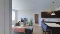 This well-designed single-floor residence showcases a clean, modern aesthetic with an open-concept layout that seamlessly connects the living and dining areas. The living space features light hardwood flooring that runs throughout, creating a warm and cohesive atmosphere. Neutral white walls and smooth ceilings contribute to the bright, airy feel of the interior, while large windows allow natural light to fill the space.

The living area provides a comfortable setting, anchored by a contemporary media console and accented with lush potted plants that add a touch of greenery to the space. A cozy seating arrangement with a patterned throw rug defines the lounge zone, making it an inviting area for relaxation.

Adjacent to the living space, the dining and kitchen area features dark-toned cabinetry with a contrasting light countertop, paired with modern bar-style seating. The open sightlines between the kitchen and living area enhance the sense of spaciousness. A hallway visible from the main area leads to additional rooms, suggesting a thoughtful and functional floor plan. Overall, the space strikes a balance between modern design and comfortable everyday living.