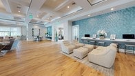 This elegantly designed single-floor amenity space offers a seamless blend of social, recreational, and professional environments, all unified by warm hardwood flooring, coffered ceilings, and a refined contemporary aesthetic.

The expansive main lounge serves as the heart of the space, anchored by a striking marble-surround fireplace with a built-in aquarium feature, a large flat-screen TV, and generous sectional seating arranged atop a textured area rug. Oversized windows and glass doors flood the room with natural light while offering views of the outdoor surroundings, including lush palm trees.

Adjacent to the lounge, a spacious recreation area features a full-sized billiards table set beneath a modern pendant chandelier, complemented by a vibrant teal accent rug. A reception and lounge zone with tufted seating and a bold blue mosaic tile feature wall adds visual drama and provides a welcoming check-in area with workstations.

The fully equipped catering kitchen boasts dual runs of dark cabinetry, bright quartz countertops, stainless appliances, and pendant lighting, with direct access to the outdoors through sheer-draped glass doors.

Two dedicated conference rooms provide professional meeting spaces, each furnished with a central conference table, executive seating, a wall-mounted display, and distinctive sculptural overhead lighting. Intimate nook seating areas with curated wall art offer quiet retreat spots throughout the space.