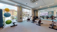 Located at 1401 King in Bridgeport, Connecticut, this well-appointed fitness center offers a comprehensive exercise environment designed to support a variety of workout routines. The space is laid out on a single level and features a clean, contemporary aesthetic with neutral-toned carpet flooring, recessed lighting, and a dropped ceiling with integrated HVAC units.

The cycling area is a focal point of the room, equipped with multiple stationary spin bikes arranged to face a large wall-mounted display screen, ideal for guided or on-demand cycling sessions. A full-length mirror spans one wall, visually expanding the space and allowing users to monitor their form during workouts.

A dedicated functional training zone includes TRX suspension straps anchored to the ceiling, a collection of kettlebells in varying weights, a rack of dumbbells, stability and medicine balls with an instructional display rack, and a flat workout bench. This area supports strength, balance, and mobility training.

Floor-to-ceiling glass doors and oversized windows provide abundant natural light and direct views of an outdoor pool and courtyard area beyond. The glass entry door features a colorful art piece in the adjacent vestibule, contributing to the space's modern, welcoming character. The overall ambiance is bright, clean, and functionally organized for diverse fitness needs.