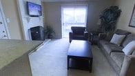 This single-floor space centers around a welcoming living area designed for both comfort and functionality. The room features a classic fireplace with a tiled surround and hearth, serving as a natural focal point along one wall, complemented by a wall-mounted flat-screen television above. Soft, neutral-toned carpeting extends throughout the living area, adding warmth underfoot. The seating arrangement includes a plush sofa and a dark leather armchair, anchored by a dark wood coffee table with a lower storage shelf — a practical and stylish centerpiece. A sliding glass door fitted with vertical blinds fills the space with abundant natural light and provides direct access to an outdoor area, offering views of the surrounding exterior. Flanking the door, a tall potted plant adds a refreshing natural accent to the room. On the opposite side, a side table with a lamp and an additional seating option create a cozy reading nook. The neutral beige walls and understated décor lend the space a calm, inviting ambiance, making it a versatile and comfortable environment suited for everyday living and relaxed entertaining.