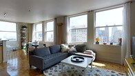 This single-floor residence offers an open-concept layout defined by abundant natural light and sweeping urban skyline views. Large double-hung windows line the living area, framing impressive cityscape panoramas and drawing in generous sunlight throughout the day. The smooth white ceilings complement the warm-toned parquet wood flooring, which runs throughout the space and adds a classic, textural contrast to the contemporary furnishings.

The living area serves as the central hub of the home, anchored by a large sectional sofa and a white coffee table atop a patterned area rug. Decorative geometric star wall accents add an artistic, modern touch to the space. A flat-screen television sits atop a bold yellow media console, contributing a vibrant pop of color.

The open layout seamlessly connects the living area to a dining space and kitchen, which features a peninsula counter with bar seating — ideal for casual dining. The kitchen maintains a functional, clean aesthetic that integrates naturally into the broader living space.

Multiple window clusters throughout the unit ensure consistent natural light from several orientations, reinforcing the airy, elevated feel of this well-appointed urban residence.