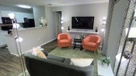 This single-floor space showcases a thoughtfully designed living area that balances comfort and contemporary style. The living room serves as the central gathering space, featuring dark hardwood-style flooring that grounds the room and contrasts nicely with the light gray walls. A wall-mounted flat-screen television is positioned on a minimalist floating shelf, flanked by tall vertical accent lamps that provide warm ambient lighting on both sides.

Two coral-toned barrel accent chairs are arranged symmetrically around a sleek black side table, creating an inviting conversational seating area. A gray sofa anchors the foreground, while a green patterned area rug ties the seating arrangement together and adds a vibrant pop of color to the space.

A pass-through opening in the wall connects the living area to an adjacent space, likely a kitchen or utility area, enhancing the open feel of the layout. Decorative touches, including small potted plants, a globe accent piece, and layered throw pillows, add personality throughout. A doorway visible in the background suggests additional rooms beyond the main living area.