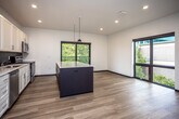 This single-floor space showcases a thoughtfully designed, contemporary layout that blends style and functionality. The open-concept kitchen and living area serves as the heart of the unit, highlighted by a striking two-tone center island with a crisp white countertop and dark navy base, complemented by pendant lighting overhead. White shaker-style cabinetry lines the kitchen walls, paired with a bold dark tile backsplash and stainless steel appliances, including a refrigerator, range, microwave, and dishwasher. Warm wood-tone flooring flows consistently throughout, tying the spaces together seamlessly.

Large black-framed windows in the kitchen and living areas invite abundant natural light and provide pleasant views of surrounding greenery. The bedrooms are well-proportioned, featuring the same warm flooring, clean white walls, dark-trimmed doors, and windows that continue the theme of natural light. A dedicated bedroom offers direct access to the bathroom, which features a dark-toned vanity with a white countertop, white tile flooring, and a full tub-shower combination with white subway tile surround. The overall design palette — neutral whites, warm wood tones, and bold dark accents — creates a clean, modern ambiance throughout the entire space.