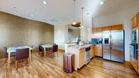 Located at 1245 E Prairie Brook Dr in Palatine, IL, this single-floor community space is thoughtfully designed to accommodate a variety of activities, from casual gatherings to structured meetings. Rich wood-look flooring runs throughout, creating a warm and cohesive aesthetic across all areas.

The kitchen is a standout feature, outfitted with light maple cabinetry, granite countertops, and a full suite of stainless steel appliances including a side-by-side refrigerator with ice and water dispenser, double wall ovens, a built-in microwave, and a dishwasher. A central island with a tiled base provides additional prep space and casual seating, while elegant pendant lighting hangs overhead. The soaring ceilings amplify the sense of openness throughout this area.

Adjacent to the kitchen, a dining and meeting area is defined by a striking curved mosaic tile accent wall in warm gold tones. The space accommodates multiple tables with comfortable upholstered seating, and a workstation with a desktop computer is available along the wall.

The lounge area exudes a refined, residential feel, anchored by a built-in entertainment center crafted from light wood with detailed millwork, housing a fireplace below a mounted television. Crisp white sofas and carefully chosen accent pieces create an inviting atmosphere, with natural light filtering in through large windows nearby.