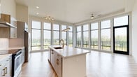 This contemporary single-floor residence is defined by its striking open-plan living and kitchen area, where an expansive arrangement of floor-to-ceiling black-framed windows floods the space with natural light and frames sweeping outdoor views. The herringbone-patterned wood flooring flows throughout, adding warmth and visual interest to the bright, neutral interior palette.

The kitchen serves as the heart of the space, anchored by a generous waterfall-edge island finished in white quartz with an integrated copper-toned sink and gold hardware. Two-tone cabinetry in light gray and natural wood tones lines the perimeter, complemented by stainless steel appliances, a wall-mounted range hood, a subway tile backsplash, and a dedicated wine cooler. Pendant and track lighting complete the refined aesthetic.

The bedroom offers a calm retreat with light hardwood flooring and a well-positioned window overlooking greenery. The primary bathroom features large-format tile, a dual-sink vanity with quartz countertops, a soaking tub, and direct access to a walk-in closet with built-in shelving.

A dedicated in-unit laundry closet houses a full-size Whirlpool front-loading washer and dryer, concealed behind louvered bi-fold doors, with overhead shelving for added storage.