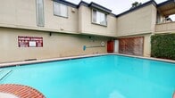 This residential complex in Santa Rosa, CA, is characterized by its well-maintained communal swimming pool, which serves as the focal point of the property. The pool area is enclosed by two-story buildings with a uniform beige exterior and distinctive navy blue trim, creating a cohesive architectural aesthetic.

The swimming pool is a standout feature, boasting a curved shape with a vibrant turquoise hue. It's bordered by a decorative brick coping in a contrasting reddish tone, adding a touch of sophistication to the design. The pool area is equipped with safety features, including handrails and clear signage displaying pool rules and hours.

Surrounding the pool, concrete pathways provide easy access for residents. The landscaping incorporates lush greenery, with ivy-covered walls and strategically placed plants adding natural beauty to the space. Comfortable lounge chairs are available poolside for relaxation.

The buildings themselves feature a mix of window styles, including some larger picture windows, allowing for natural light in the units. Privacy lattice and wooden fencing separate individual outdoor spaces. The overall design suggests a focus on creating a pleasant, community-oriented living environment with a balance of shared and private spaces.